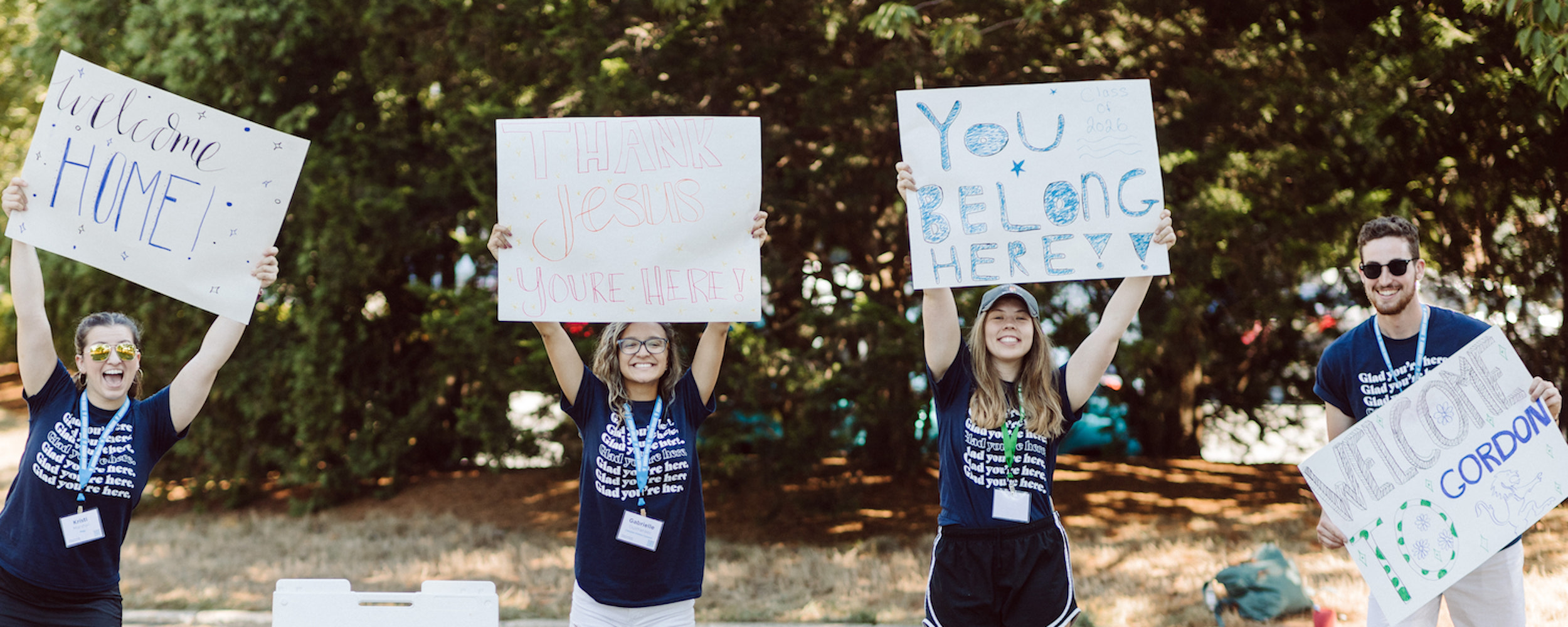 Four orientation leaders at Gordon College welcoming new students on campus with signs.