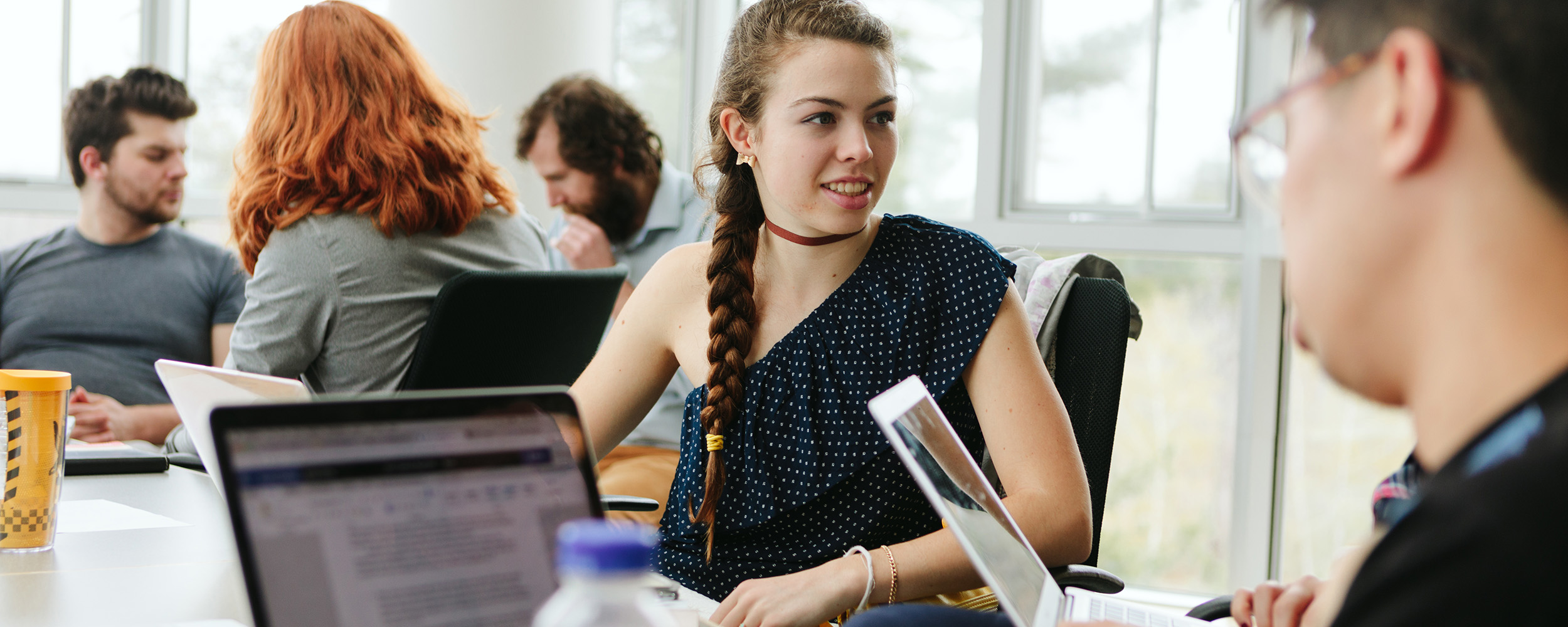 students in college for journalism sitting in small groups around a classroom writing articles on laptops