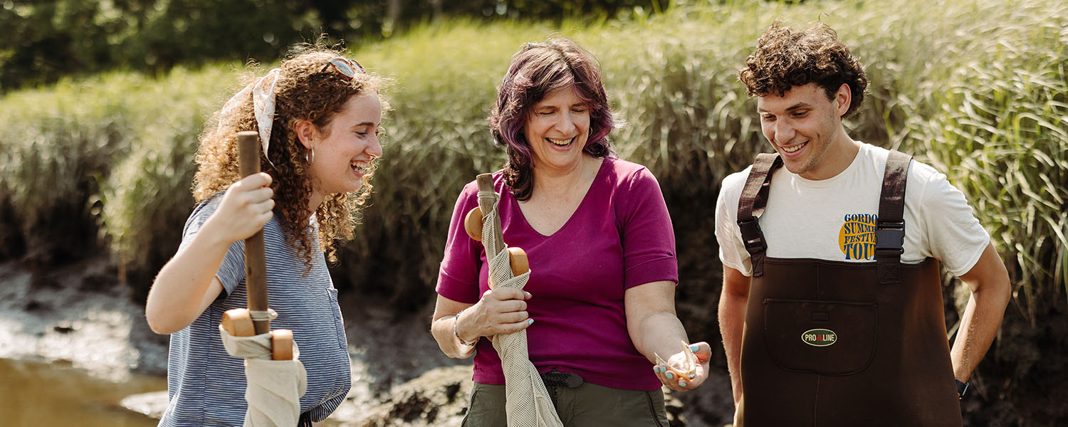 Two marine biology students at Gordon College laughing and learning with their professor while exploring a local marsh