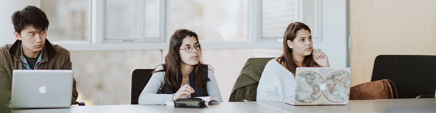 Three students in a classroom with their laptops engaged in discussion